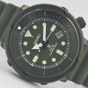 May include: A Seiko solar diver's watch with a dark olive green dial and a matching olive green silicone strap. The watch features a black rotating bezel, luminous hour markers, white hands, and a date window at the three o'clock position.