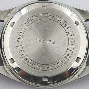 May include: Close-up of the back of a stainless steel watch with a black leather strap. The watch face is silver with the words "Shock Protected", "Stainless Steel", and "Water Dust Protected" around the edge. The number "140172" is in the center.