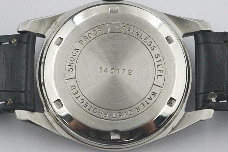 May include: Close-up of the back of a stainless steel watch with a black leather strap. The watch face is silver with the words "Shock Protected", "Stainless Steel", and "Water Dust Protected" around the edge. The number "140172" is in the center.