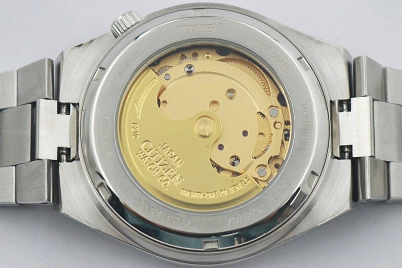 May include: Close-up of the back of a Citizen watch, showcasing the intricate internal mechanism. The watch has a silver-tone stainless steel band and case. The visible movement features gold-colored gears and a rotor, with text that reads "Citizen Watch Co."