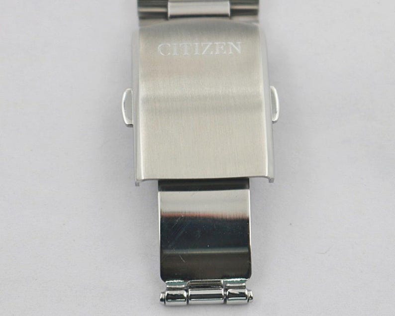 May include: A stainless steel watch clasp with the brand name "CITIZEN" engraved on the top. The clasp is rectangular with rounded edges and a hinged section below. The watch band is attached to the clasp.