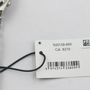 May include: A close-up of a silver wristwatch with a pink watch face and a metal band. A white tag attached to the watch displays the text "NJ0158-89X Cal. 8210" and a barcode.