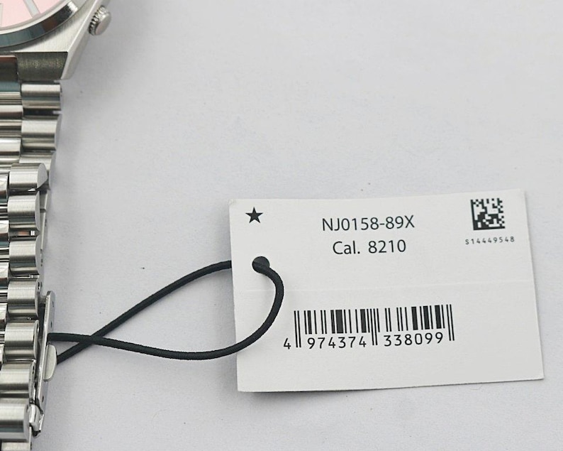 May include: A close-up of a silver wristwatch with a pink watch face and a metal band. A white tag attached to the watch displays the text "NJ0158-89X Cal. 8210" and a barcode.