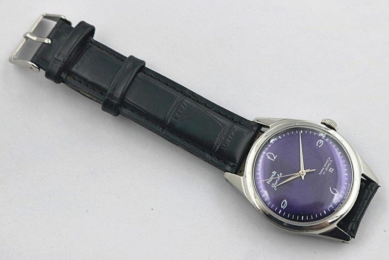 May include: A vintage wristwatch with a dark purple face and silver hands and casing. The watch has a black leather band with a silver buckle. The watch face has white numerals and text.