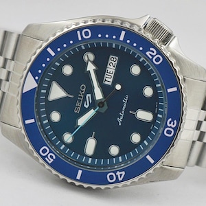 Puede incluir: Un reloj automático Seiko 5 de acero inoxidable con esfera y bisel azules. El reloj presenta marcadores de hora blancos, una pantalla de día y fecha y una pulsera plateada. El bisel tiene marcas blancas y un fondo azul. Un reloj clásico.