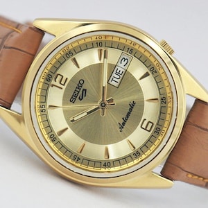 May include: A gold-tone Seiko 5 automatic wristwatch with a sunray gold dial, gold-tone hands and indices, and a brown leather strap with a crocodile-embossed texture. The dial features the text Seiko 5, Automatic, and a day-date window showing TUE 3.