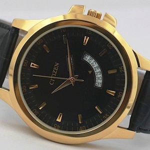 May include: A gold-toned Citizen watch with a black face and a black leather band. The watch features gold hands and markers, a date display, and the brand name "CITIZEN". The watch band is approximately 9 inches long.