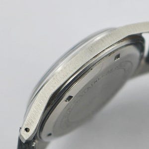 May include: Close-up of a silver-toned wristwatch with a black leather strap. The watch has a round face and a brushed metal finish. The watch case is stainless steel. The watch is angled, showing the side and back of the watch.