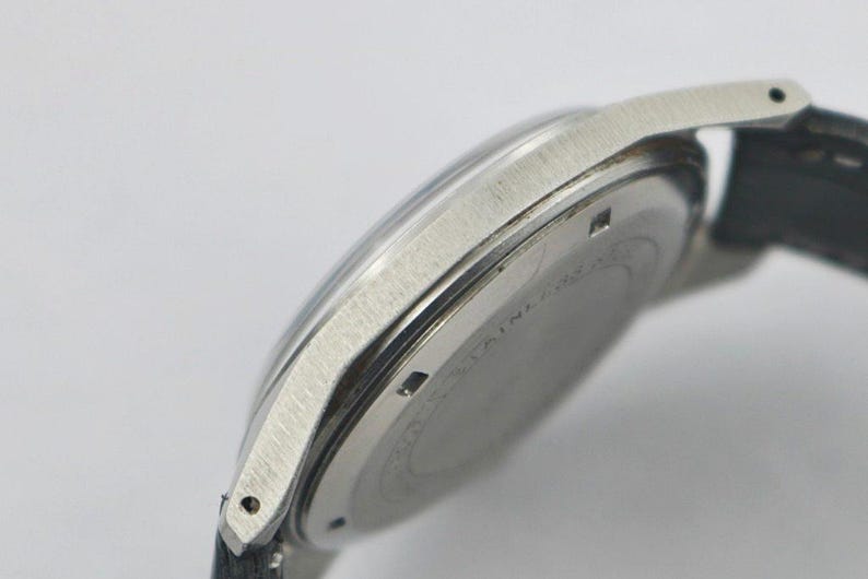 May include: Close-up of a silver-toned wristwatch with a black leather strap. The watch has a round face and a brushed metal finish. The watch case is stainless steel. The watch is angled, showing the side and back of the watch.