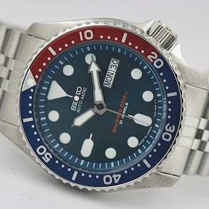 May include: A stainless steel automatic wristwatch with a dark blue dial, featuring luminous circular and rectangular hour markers, a day-date window, and a red and blue rotating bezel. The watch includes a silver-tone metal link bracelet.