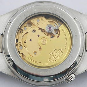 May include: Close-up of the back of a silver Citizen watch, showcasing the intricate gold-toned mechanical movement. The watch has a stainless steel band and a clear glass back, revealing the inner workings. The words "JAPAN CITIZEN WATCH CO." are visible.