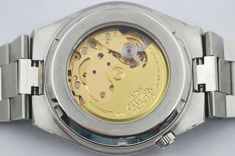 May include: Close-up of the back of a silver Citizen watch, showcasing the intricate gold-toned mechanical movement. The watch has a stainless steel band and a clear glass back, revealing the inner workings. The words "JAPAN CITIZEN WATCH CO." are visible.