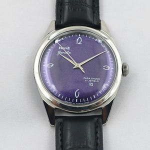 May include: A vintage wristwatch with a silver-tone case and a deep purple dial. The watch face features silver hands and hour markers, with the text "HMT Janata" and "Para Shock 17 Jewels". The watch has a black leather band.