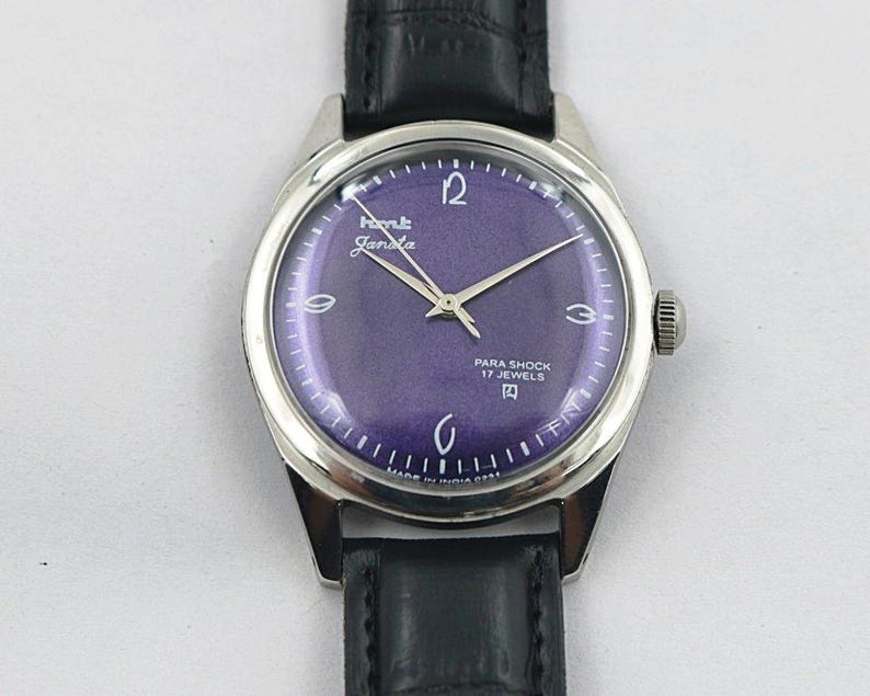 May include: A vintage wristwatch with a silver-tone case and a deep purple dial. The watch face features silver hands and hour markers, with the text "HMT Janata" and "Para Shock 17 Jewels". The watch has a black leather band.