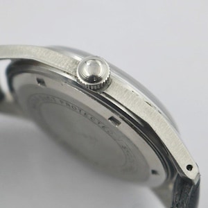 May include: Close-up of a silver-toned wristwatch with a black leather band. The watch features a round face, a textured bezel, and a crown. The watch's back has engraved text. The watch is a classic timepiece.
