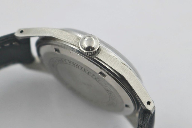 May include: Close-up of a silver-toned wristwatch with a black leather band. The watch features a round face, a textured bezel, and a crown. The watch's back has engraved text. The watch is a classic timepiece.