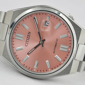 May include: A Citizen automatic wristwatch with a salmon-colored dial and silver-tone hands and markers. The watch features a date window at the 3 o'clock position and a stainless steel band. The watch face is approximately 1.5 inches in diameter.