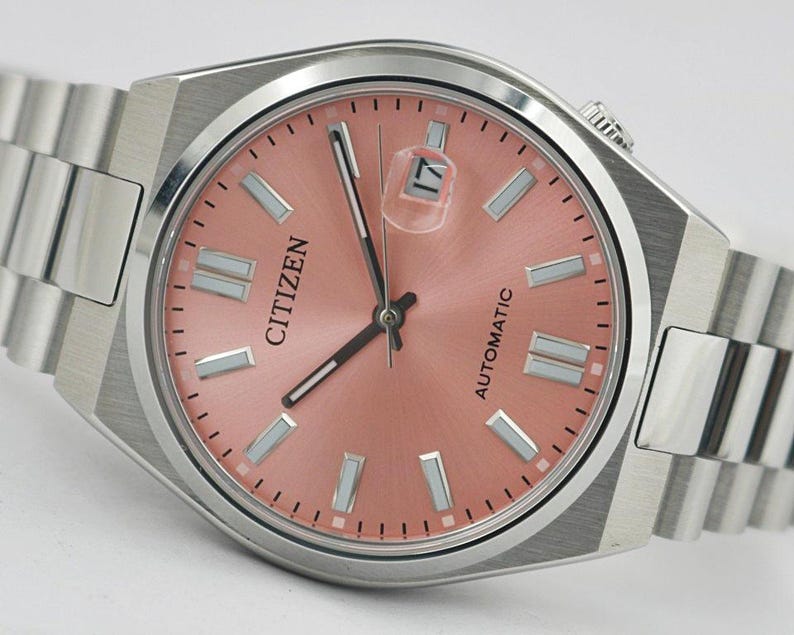 May include: A Citizen automatic wristwatch with a salmon-colored dial and silver-tone hands and markers. The watch features a date window at the 3 o'clock position and a stainless steel band. The watch face is approximately 1.5 inches in diameter.