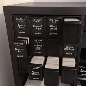 Könnte beinhalten: Ein schwarzes Aufbewahrungsregal mit mehreren Fächern, die jeweils mit weißem Text beschriftet sind. Die Etiketten lauten "One Piece" gefolgt von verschiedenen Episodentiteln. Die Fächer sind teilweise geöffnet und zeigen Kartenstapel.