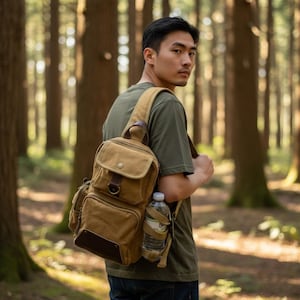 Op de afbeelding: Een bruin canvas sling bag met meerdere vakken en een waterfleshouder. De tas heeft bruine leren accenten en wordt over de schouder gedragen. De persoon draagt een groen t-shirt en een blauwe jeans.