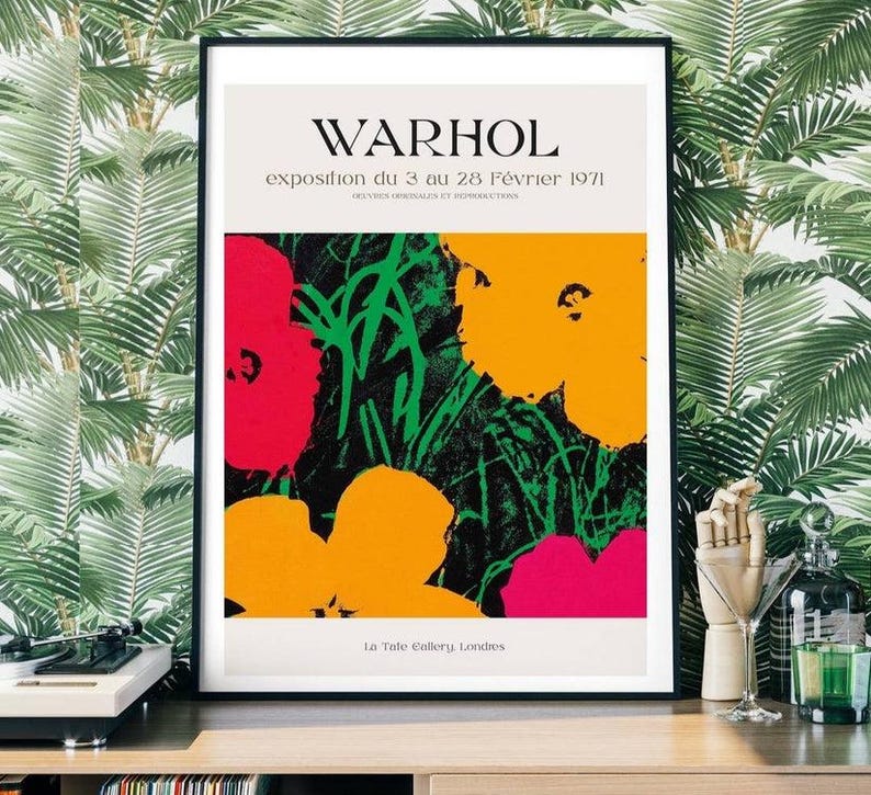 May include: A framed art print featuring a colorful floral design in red, yellow, and pink against a black and green background. The print includes the text "WARHOL" and exhibition details in French. The artwork is displayed in a black frame.
