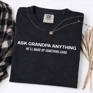 May include: Black t-shirt with the text "ASK GRANDPA ANYTHING HE'LL MAKE UP SOMETHING GOOD" in white. A silver necklace with a small pendant rests on the shirt. A plaid shirt and dried plants are in the background.