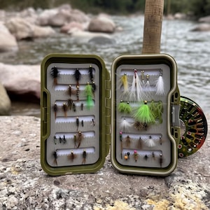May include: An olive green plastic fly fishing tackle box open to reveal two foam inserts holding an assortment of artificial fishing flies in various colours including black, white, green, and brown, with a cork handle and reel visible behind.