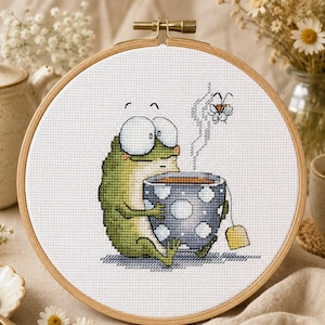 May include: A cross-stitch embroidery in a round wooden hoop featuring a green frog with large white eyes holding a blue and white polka-dot mug of steaming tea. A small fly is depicted above the mug, and a yellow tea bag tag hangs from the side.