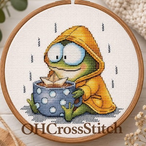 May include: A cross-stitch embroidery in a wooden hoop featuring a green frog wearing a yellow hooded raincoat, holding a blue polka-dot mug with a paper boat inside, set against a background of falling rain and the text OHCrossStitch.