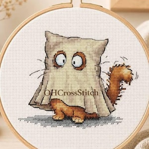 以下が含まれることがあります： 木製の丸い刺繍枠に、お化けの仮装のようにベージュの枕カバーを頭から被ったオレンジ色の猫のクロスステッチ模様が施されています。猫は大きな目とふさふさした尻尾をしています。中央にはOHCrossStitchという文字が印刷されています。