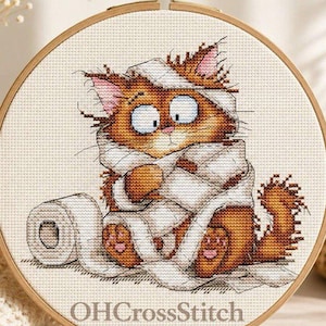 May include: A circular wooden embroidery hoop containing a cross-stitch pattern of a fluffy orange cat wrapped in white toilet paper. The cat has wide, surprised eyes and pink paw pads. The text OHCrossStitch is printed in black at the bottom.