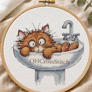 May include: A circular wooden embroidery hoop containing a cross-stitch pattern of a fluffy orange cat with wide eyes sitting inside a white bathroom sink with a silver faucet. The text OHCrossStitch is printed in brown across the sink basin.