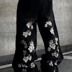 Może przedstawiać: Czarne spodnie z szerokimi nogawkami z białym haftem kwiatowym po bokach. Spodnie mają klasyczny krój z pięcioma kieszeniami i są noszone z białymi trampkami. Haft dodaje dekoracyjny element do swobodnego stylu.
