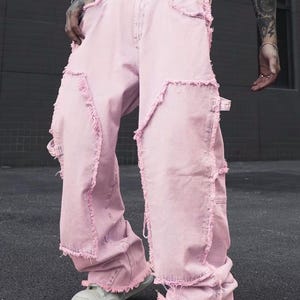 May include: Light pink denim jeans with distressed edges and a wide leg fit. The jeans feature a white belt with silver studs and are paired with white sneakers. A modern take on a classic style.