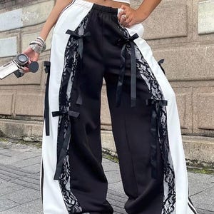 May include: Black and white wide-leg pants with lace and ribbon accents. The pants feature a split design with a black panel on one side and a white panel on the other. Black ribbons are tied along the sides, and black lace trim adds detail.