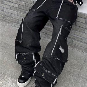 May include: Black denim cargo pants with white stitching and distressed details. The pants feature multiple pockets and a wide leg design. The model is wearing black and white sneakers.