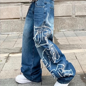Peut inclure: Jean bleu avec lettrage blanc vieilli sur les jambes. Le jean a une coupe large et est porté avec des baskets blanches. Le jean est porté avec une ceinture cloutée.