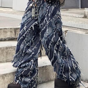 Peut inclure: Jean déchiré bleu foncé avec un design effiloché unique. Le jean présente des rayures verticales et des bords effilochés blancs. Le pantalon large est associé à des bottes à plateforme noires, pour un look audacieux.