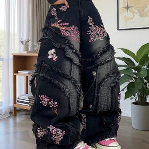 May include: Black denim jeans with floral embroidery in pink and white. The jeans have a wide leg design with frayed edges and are paired with pink canvas sneakers.