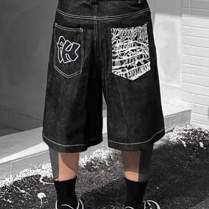 May include: Black denim shorts with white stitching and large back pockets. The left pocket features a white graphic of the letters "fk", while the right pocket has a white, intricate design. The shorts are worn with black socks and black and white sneakers.