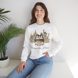 Può includere: Felpa bianca con grafica di due pinguini che si abbracciano davanti a un castello. I pinguini hanno cuori con i colori delle bandiere ucraina e tedesca. Il testo recita "Dva sercya, eine Heimat".