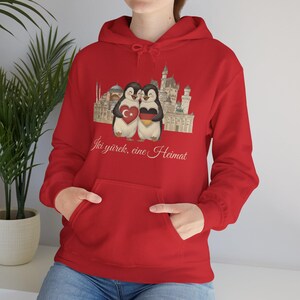 Può includere: Felpa con cappuccio rossa con una grafica di due pinguini che tengono un cuore con bandiere turche e tedesche, con uno sfondo di paesaggio urbano. Il testo "Iki yürek, eine Heimat" è stampato sotto. La felpa ha una tasca frontale.