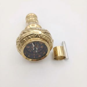 May include: A gold-toned brass handle featuring an integrated circular compass with a black face and white directional markings. The handle has an ornate, textured surface. Beside it lies a small, detached cylindrical brass collar and a metal pin.