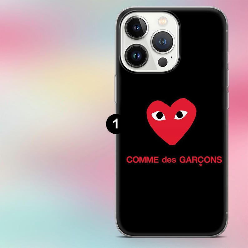 K&ouml;nnte beinhalten: Schwarze Handyh&uuml;lle mit rotem Herz-Logo und dem Text "COMME des GAR&Ccedil;ONS". Die H&uuml;lle hat einen transparenten Rand und einen Kameraausschnitt.