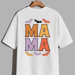 Pode incluir: Uma t-shirt branca de manga curta com um gráfico nas costas que apresenta a palavra MAMA em letras grandes com textura de madeira em tons de laranja, amarelo e roxo. O texto é rodeado por pequenas ilustrações coloridas de morcegos.