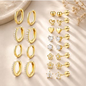 May include: A collection of gold-toned earrings including four pairs of hoop earrings and seven pairs of stud earrings. The hoops feature plain and crystal-encrusted designs. The studs include spheres, hearts, flowers, and crystal-set shapes.