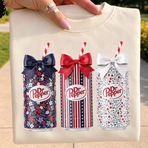 Könnte beinhalten: Ein cremefarbenes Sweatshirt mit Rundhalsausschnitt und einer Grafik von drei Dr Pepper-Dosen. Jede Dose ist mit einer Schleife und einem rot-weiß gestreiften Strohhalm versehen. Die Dosen zeigen florale Muster sowie rot-weiß-blaue Streifen.