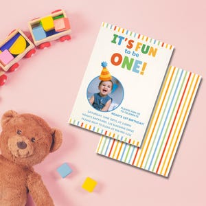 Puede incluir: Una invitaci&oacute;n de cumplea&ntilde;os con el texto "IT'S FUN to be ONE!" presenta una foto de un beb&eacute; con un gorro de fiesta. La invitaci&oacute;n incluye detalles del evento y un dise&ntilde;o de rayas coloridas en la parte posterior. Un oso de peluche y bloques de juguete de madera est&aacute;n en el fondo.