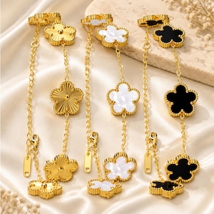 May include: Three gold-toned chain bracelets featuring flower-shaped charms. The first bracelet has textured gold flowers, the second has white floral charms, and the third has black floral charms. Each bracelet includes a lobster clasp and a small tag.