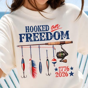 以下が含まれることがあります： 青と赤で「HOOKED on FREEDOM」の文字がプリントされたクリーム色のTシャツ。グラフィックには、リール付きの釣り竿と、羽や浮きを含む5つのぶら下がったルアー、そして青い星を伴う「1776」と「2026」という年号が描かれています。